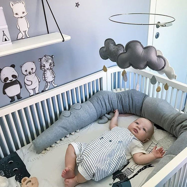 A cute baby lying in a crib surrounded by gray cartoon baby bedding, featuring a playful bumper pillow from Baby Bliss Haven, perfect for infant safety and comfort.